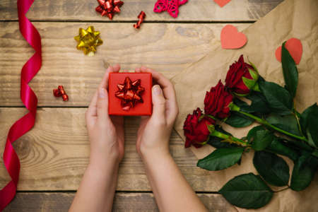 Top view, female hands holding a red gift box. Festive atmosphere, red roses, satin ribbon and bows on a wooden background.の写真素材