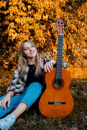 A young girl is playing guitar in the park. autumn yellow leaves. Hobby or study. A girl in jeans and a plain shirt. student and teenager. Generation Zの写真素材