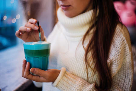 A young woman holds a blue cocktail in her hands. A girl in a white warm sweater. Cozy cafe.の写真素材