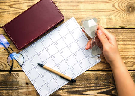 A blank sheet of notepad and a calendar for planning for a month or a year. Glasses, notepad and pen on a wooden background. Workspace. A woman's hand holds an hourglass.の写真素材