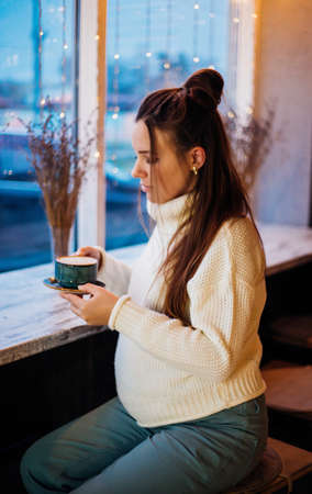 A young pregnant woman is sitting at a table in a cafe. The girl drinks hot coffee. Waiting for the birth of a child. Cozy evening.の写真素材