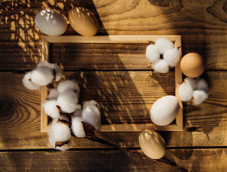 Eggs and a branch with white cotton flowers with sunny shadows on a wooden background . Twigs are...