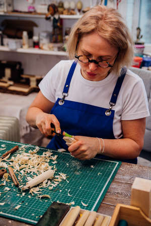 A female carpenter cuts wood with a knife in a carpentry or makeshift workshop. Dust and shavings scatter in the air.Workplace. Manufacture of wooden toys.の写真素材
