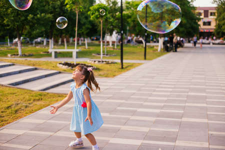 In a public park, a little girl blows and catches soap bubbles. The child is playing and laughing. The bubbles shimmer in the sun and fly in different directions.の写真素材