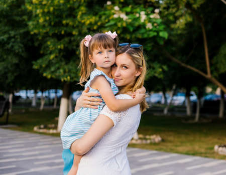 A young mother holds her little daughter in her arms. A strong loving hug. Family walk in the park in summer.の写真素材