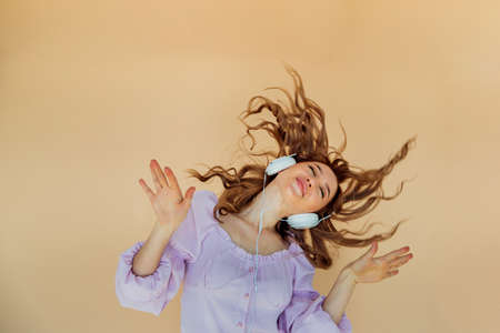 A beautiful young woman listens to music with headphones and sings on a beige background. A charming girl with headphones dancing with her hair and eyes closed.の写真素材