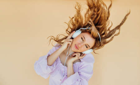 A beautiful young woman listens to music with headphones and sings on a beige background. A charming...