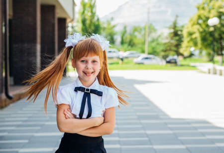 A little girl, an elementary school student, stands on the street with her hands folded. Portrait of a schoolgirl with red hair. Back to school.の写真素材