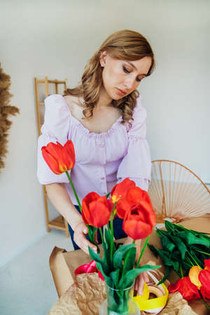 A young woman is packing a festive bouquet in wrapping paper. The florist makes an assembly with red tulips in the workshop. A woman at work, in a small business or hobby.の写真素材