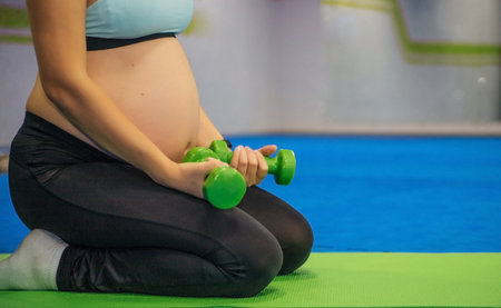 A pregnant woman does sports, holds dumbbells in her hands. sports uniform. healthcare. waiting for a child. Belly close-up. Space for text.の写真素材