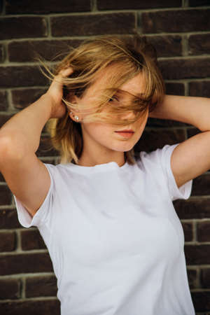 Portrait of a young beautiful girl looking into the camera against a brick wall. A fashionable teenager in casual summer clothes. Her hair is blown away by the wind and falls on her face.の写真素材