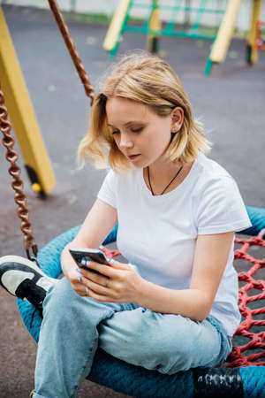 A cute teenage girl holding a phone in her hand is sitting on a swing in a modern park. A schoolgirl...