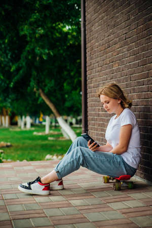 A young beautiful teenage girl is sitting on a skateboard on a beautiful summer day in the park. A girl is talking by video on a mobile phone in the open air. Chat with friends.の写真素材