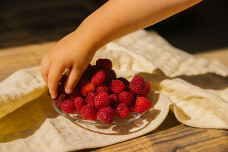 A small saucer with ripe raspberries, a bunch of summer berries on a wooden background. A child's hand takes a berry from the dishes. summer background. Juicy appetizer or dessert.の写真素材