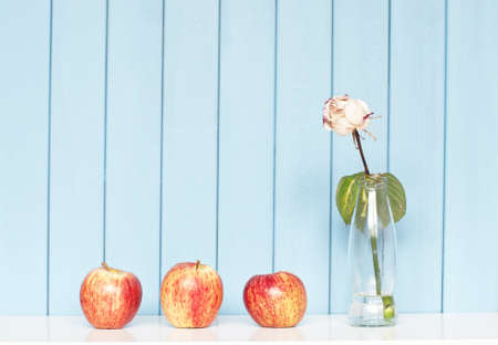 Three red apples and white rose in the bottle on the shelf on blue wooden backgroundの写真素材