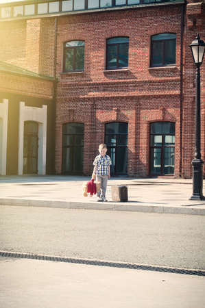 little boy with teddy bear toy and suitcase  shows forward, stands on the sunny deserted streetの写真素材