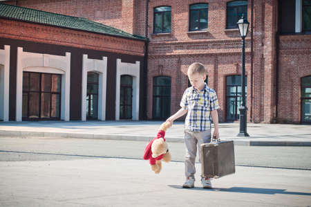 little kid is holding the suitcase and teddy bear toy and walking down the sunny street in the morningの写真素材