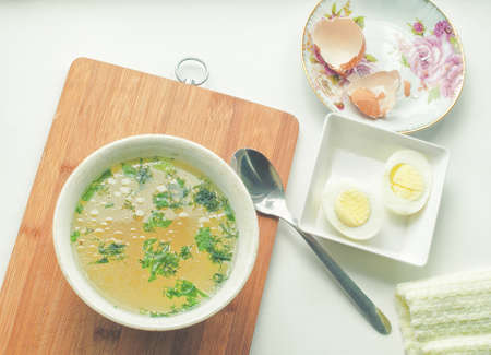 Hot chicken soup in bowl, spoon, eggs, eggshell and cloth napkin on rustic wooden tableの写真素材