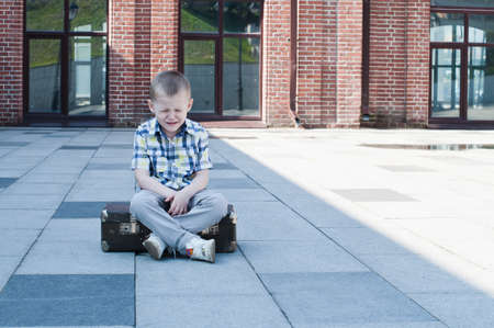 Little sad 4 years old boy is sitting on suitcase and crying on the streetの写真素材