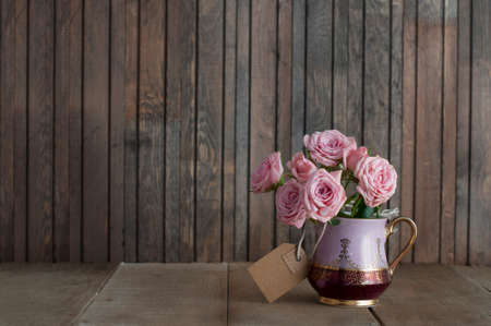 Beautiful pink roses in a vintage jug with craft paper tag on rustic wooden background