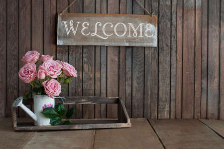 Wooden sign Welcome and Beautiful pink roses in a vintage jug on rustic wooden backgroundの写真素材