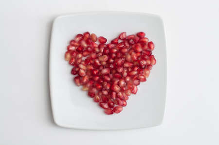 Red pomegranate juicy seeds in shape of heart in a dish on white kitchen tableの写真素材