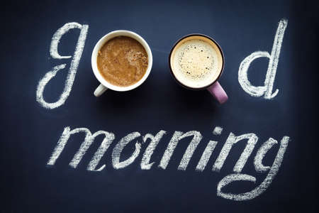 Inscription "good morning" written with chalk and two cups of coffee, dark blue backgroundの写真素材