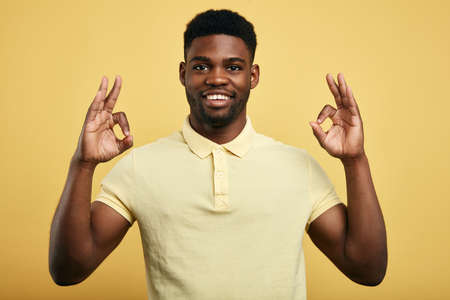positive guy in stylish T-shirt shows OK gesture with both hands on a yellow background.,...