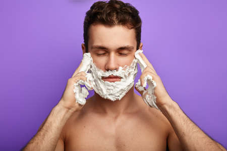 man with shaving gel on face puts fingers on temple. guy with closed eyes touching his temples and trying to concentrate on thoughts, man having headache while shaving in morning, meditation conceptの写真素材