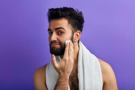 A handsome man with a towel around his neck applies , puts shaving foam on his face before shaving....