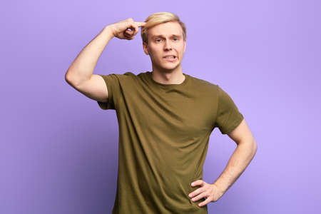 blonde frustrated angry sad man with hand on his hip points at head. close up photo. isolated violet background.guy doesnt agree with his friend. Are you mad Are your stupidの写真素材