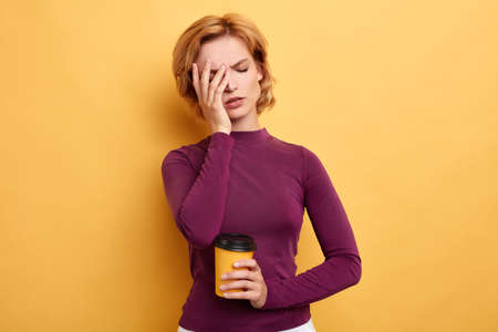 Tired sleepy beautiful woman covers half of face with palm, has sad expression, closes eyes, holding disposable cup with tea in the morning, isolated yellow background. studio shotの写真素材