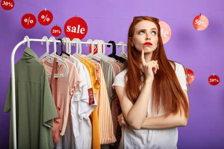 doubting beautiful young woman with finger on her chin, cheek thinking and choosing clothes isolated over blue background, girl doesnt know what clothes to buyの写真素材