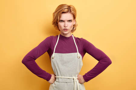 frustrated frowning housewife with hands on her hips wearing kitchen apron posing on yellow background. negative emotion and feeling. frustration and problemの写真素材