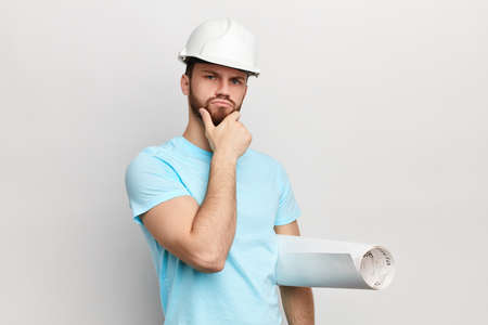 Pensive man with hand on his chin holding blueprint looking at the camera .puzzled man in hat....