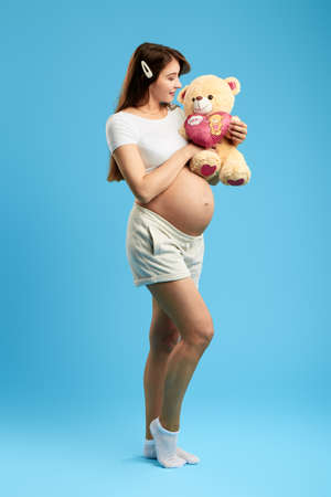 pleasant attrctive woman with a big belly olaying with teddy bear. full length photo. isolated blue background. studio shot. entertainment conceptの写真素材