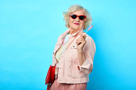Old cheerful happy lady with sunglasses holding lollipop and looking at the camera on a blue background. sweets, love, valentines day. fashion conceptの写真素材