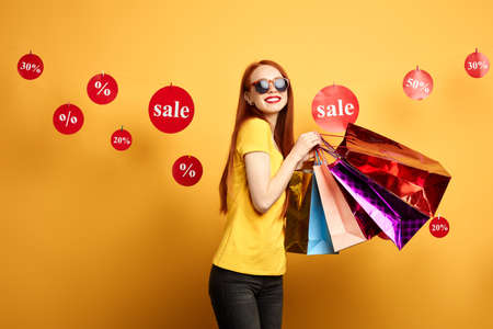 glamour charming girl in stylish T-shirt with a lot of shopping packages posing to the camera. close up portrait. free time, spare time, isolated yellow backgroundの写真素材