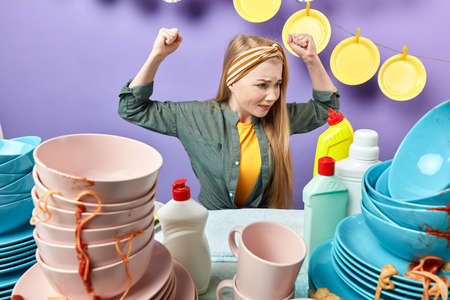 angry frustrated young woman with clenched raised fists sitting behind messy table in the kitchenの写真素材