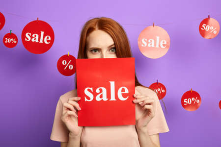 funny woman with raised eyebrow hiding behind the sale sign, isolated over blue background, studio shot.の写真素材