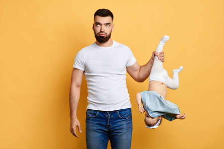 angry nervous father looking up,holding his naughty baby by leg upside down. close up photo. isolated yellow background. daddy feds up looking after his daughter. studio shotの写真素材