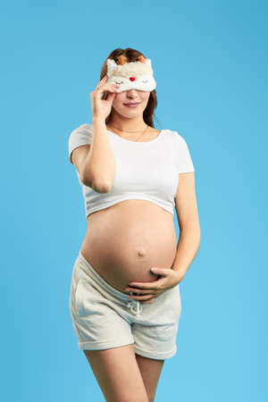 Pregnant attractive woman wearing sleeping mask on eyes posing to the camera in the morning,...