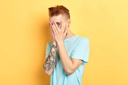 Worried stylish ginger man businessman covering face with both hands, feeling stressed out after his mistakes during the work, looking at camera through his fingersの写真素材