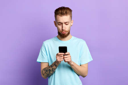 Upset stressed man with funny beard and moustache holding cellphone, received sms, message with bad news, isolated on blue background. Human face expression. emotion, feeling. close up portraitの写真素材