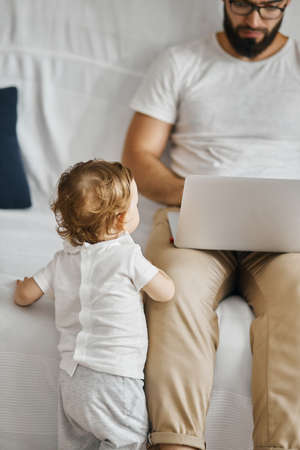 young father spending time with baby. young handsome father and cute using laptop at home, busy dad doesnt have time for his child , close up cropped photoの写真素材