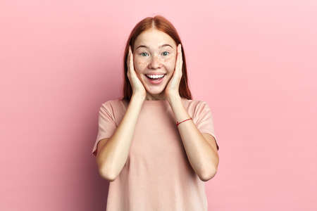 Portrait of red-headed young girl, touching cheeks with palms. Copy space. Isolated over pink...