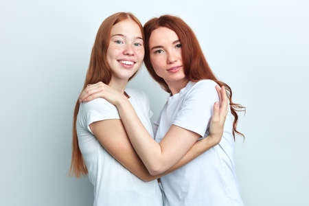 Two beautiful ginger women in casual stylish white T-shirts smiling and hugging each other isolated...