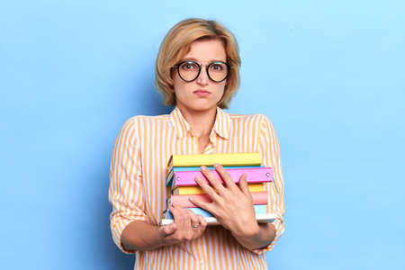 Sad unhappy young girl holding folders, getting a lot of work standing in studio blue...