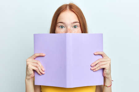 red-headed scared woman closing her face with a copy book, looking at the camera. close up portrait,...