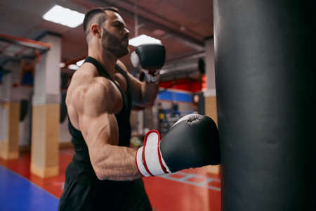 Young strong ambitious athlete working out by throwing punches at a heavy punching bag, close up...